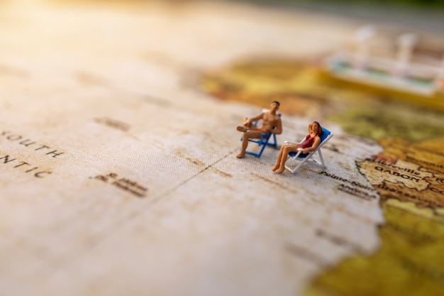 Miniature people sit on beach sunbath seats on Vintage World Map and ship, Summer Concept.