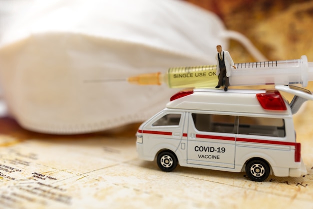 Miniature people stand on ambulance car with medical mask and Syringe of COVID-19 vaccine . Vaccine and Healthcare Medical concept.
