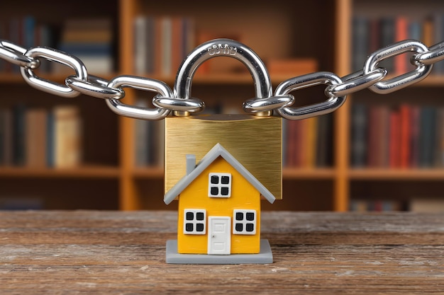 Photo miniature yellow house chained to a large metallic padlock on a wooden surface