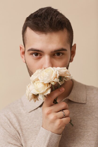 Minimal close up portrait of feminine young man holding flowers and looking at camera while posing against neutral beige background in studio