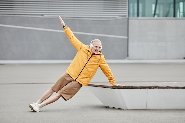 Minimal full length portrait of sportive mature man doing exercises outdoors with headphones in urba