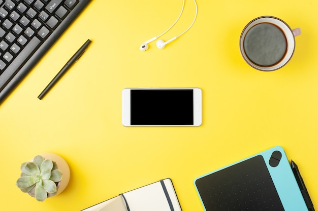 Minimal office desk. Flat lay - smartphone mock up, keyboard, earphones, tablet on yellow background. Top view. Workplace concept. Place for text.