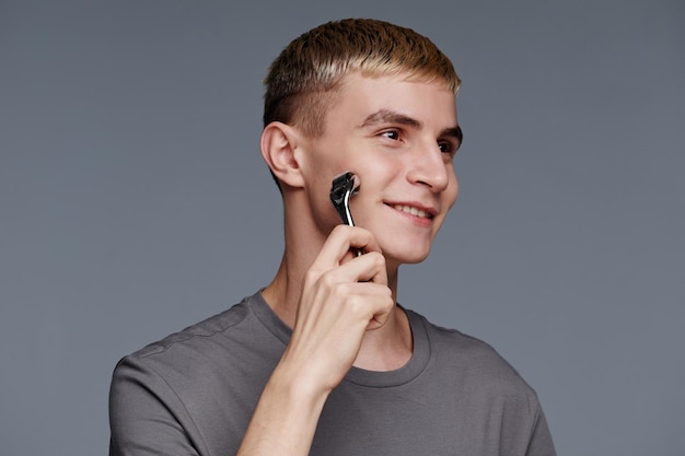 Minimal portrait of blonde young man using face massager while enjoying male skincare routine against grey background