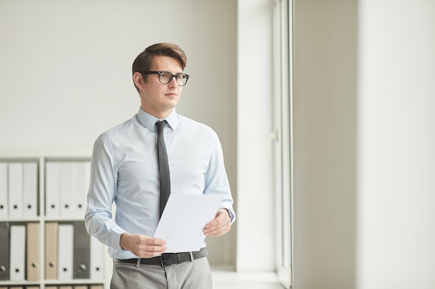 Minimal waist up portrait of young successful businessman wearing glasses and holding document while looking away standing against white wall in office, copy space