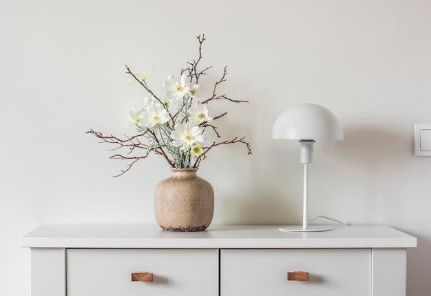 Minimalism style interior decor flower arrangement in a ceramic vase and a white metal table lamp on a white table