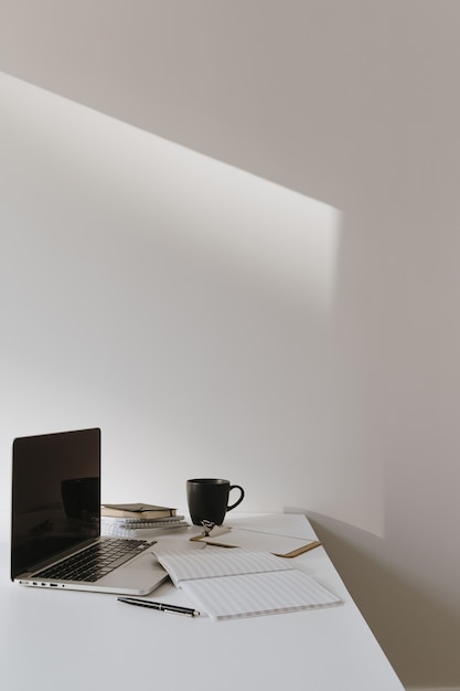 Minimalist home office desk workspace. Laptop on table with coffee cup, paper sheet, stationery against white wall