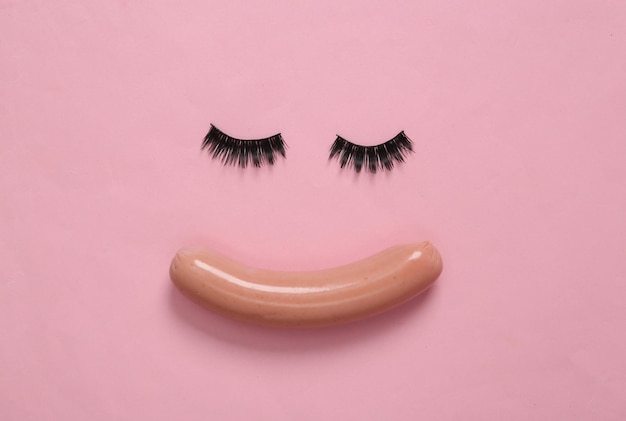 Minimalistic beauty concept False eyelashes and sausage in the shape of face on pink background