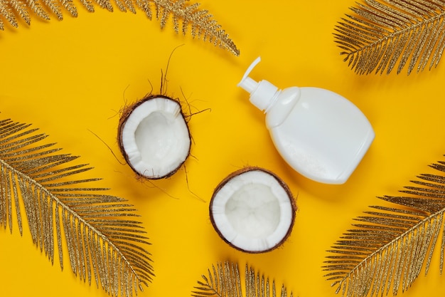 Minimalistic beauty still life. Two halves of chopped coconut and white bottle of cream with golden palm leaves on yellow background. Creative fashion concept.