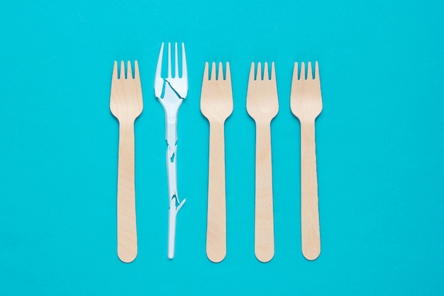 Minimalistic ecologically clean still life. Broken plastic fork among many wooden forks on blue background. Cutlery made from natural materials