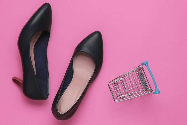 Minimalistic fashion and shopping concept. Leather high heel shoes, shopping trolley on pink pastel background. Top view