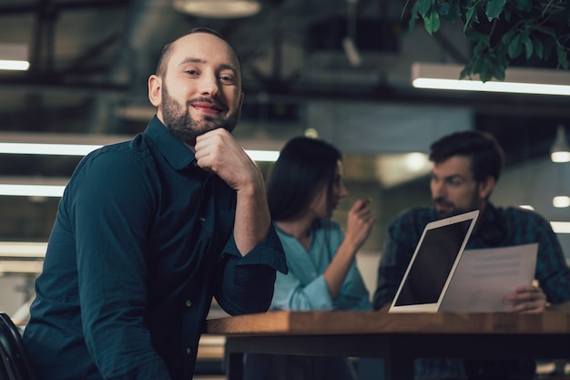 Mirthful attractive employee sitting at the table with a modern laptop and touching his chin while smiling. Two people talking on the background