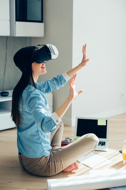 Mirthful lady sitting cross legged on the floor in front of a laptop and wearing virtual reality device