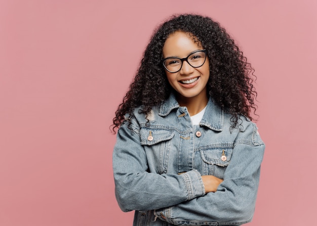 Mirthful pleasant looking female model wears optical glasses and jean jacket, keeps arms folded over chest