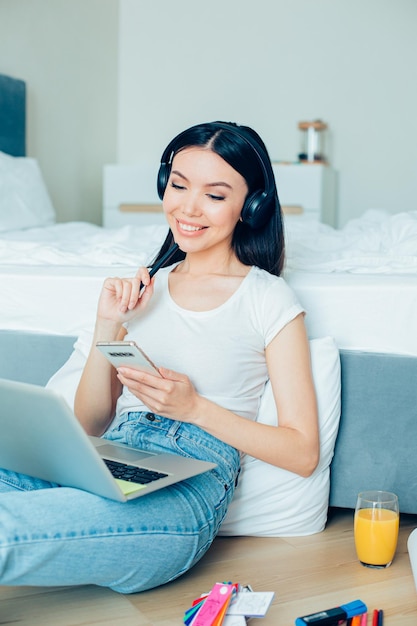 Mirthful young designer sitting on the floor with a smartphone and stationery supplies and looking at the laptop screen