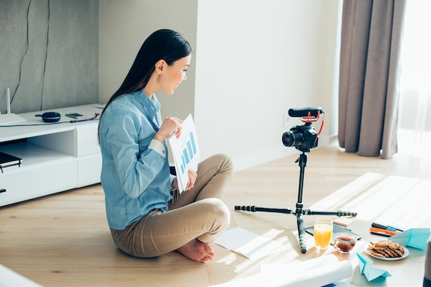 Mirthful young woman sitting barefooted with a graphic chart in her hands and smiling to a camera on tripod