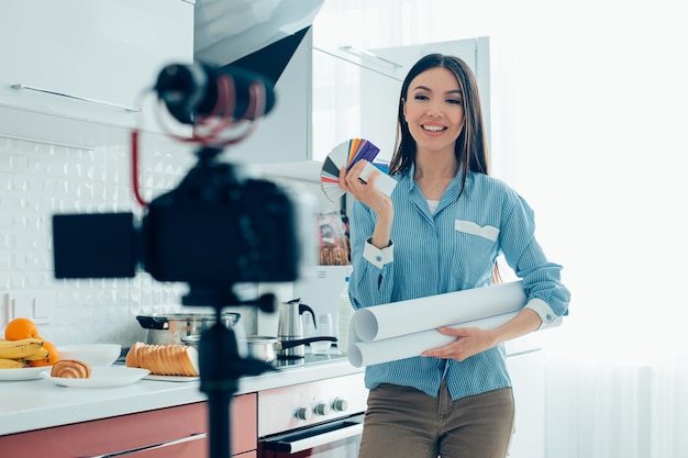 Mirthful young woman standing in the kitchen with blueprints and color palette while recording a video for a blog