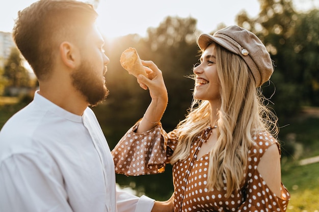 Mischievous blonde girl in beige cap tries to stain her boyfriends nose with ice cream Woman and man laughing and fooling around in park