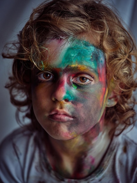 Mischievous boy with curly hair in dirty wear with colorful paint on face looking at camera while sitting in light room