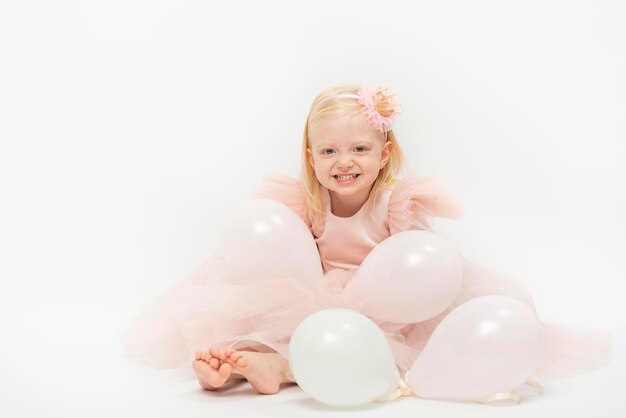 Mischievous child sits on floor in balloons Blonde girl in elegant dress and pink hoop laughs isolated on white background