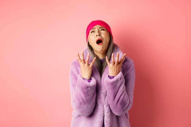 Miserable asian senior woman in stylish purple faux fur coat, pleading god and asking why, shaking hands and looking up desperate, standing over pink background