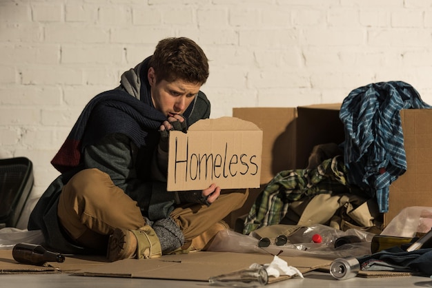 Misery man sitting on garbage dump and holding piece of cardboard with 'homeless' inscription