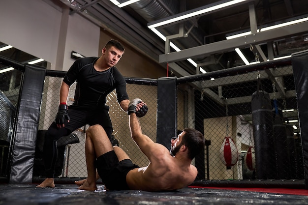 Mixed martial artists during fight, help each other, in ring at gym. Man in black wear holds out hand to shirtless male, engaged in kickboxing