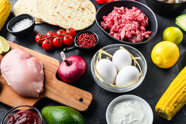 Mixed mexican food background, raw organic ingredients for tacos with chicken and beef meat, corn tortilla, salsa, chilli over black background, side  view.