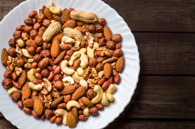 Mixed nuts in a white plate, on a dark background. Place for signature. Cashew, walnut, peanuts, hazelnuts, figs, raisins and dried apricots. 