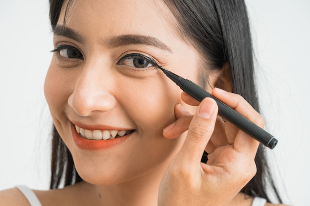 Mixed race asian woman putting eye liner on eyelid on white wall