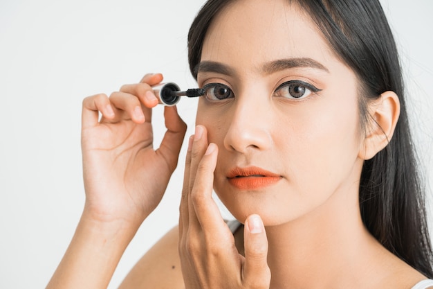 Mixed race attractive asian woman putting mascara on eyelashes on white wall