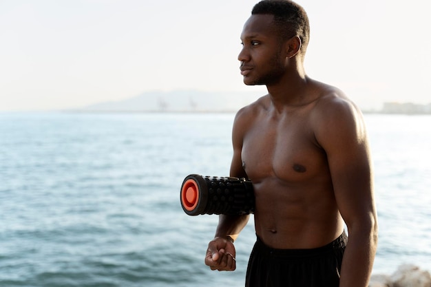 Mixed race man with naked torso standing at the seashore with yoga mat and looking away