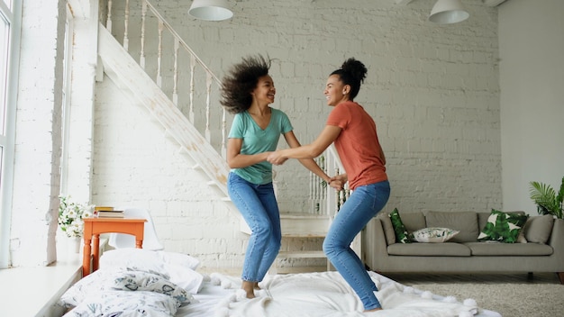 Photo mixed race young beautiful girls dancing on a bed together having fun leisure in bedroom at home