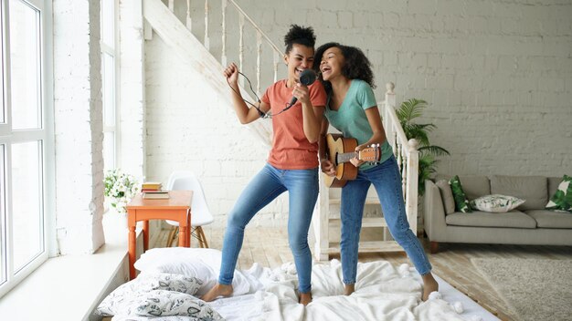 Mixed race young funny girls dance singing with hairdryer and playing acoustic guitar on a bed