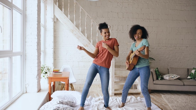 Mixed race young funny girls dancing singing and playing acoustic guitar on a bed