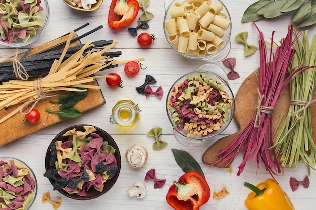 Mixed raw colorful italian pasta with various vegetables and spices on white wooden background, top view, copy space