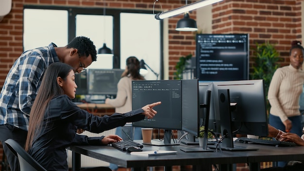 Mixed team of software developers analyzing source code pointing at screens comparing algorithm with user interface on digital tablet. Programmers working on coding group project sitting at desk.