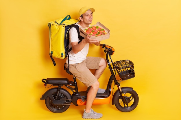 Mmm how delicious Satisfied delivery man enjoys pleasant smell from freshly baked pizza on cardboard box has good appetite transports delicious snack for clients poses over yellow background