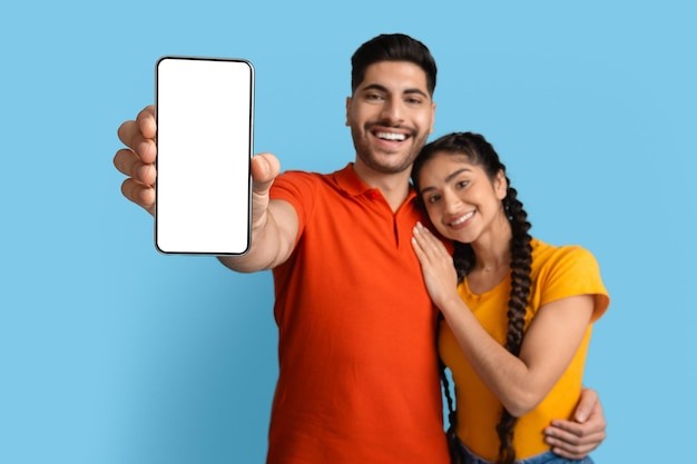 Mobile ad cheerful middle eastern couple showing big blank smartphone at camera