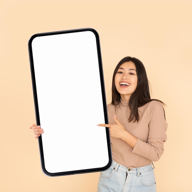 Mobile ad smiling asian woman holding and pointing at big blank smartphone