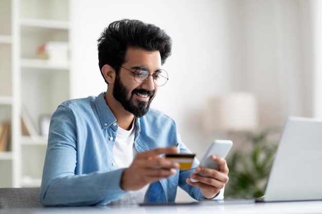 Mobile banking smiling indian man using smartphone and credit card in office