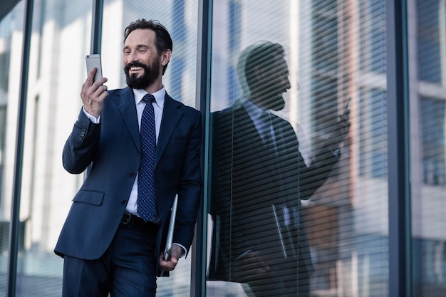 Mobile communication. Cheerful handsome businessman using his phone while standing against the wall