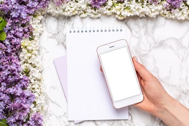 Mobile phone in hand, notebook and frame of white and lilac flowers on marble table in flat lay style. 