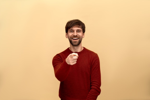 Mock, funny joke. Photo of young man with beard wearing sweater, pointing finger and toothy lough, posing against beige wall.