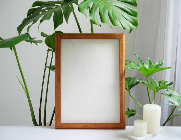 Mock up brown poster frame, gouse plant Philodendron green leaf and aroma white candles on beige table and wall background,white soft tone interior