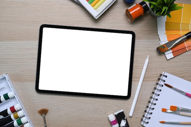 Mock up digital tablet surrounded by color swatches, paintbrushes and other various equipment on wooden background.