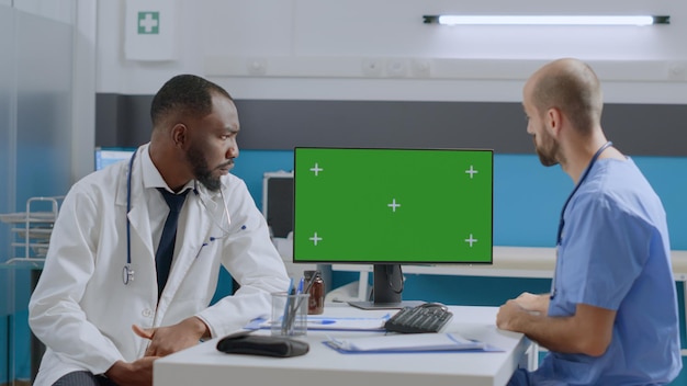 Mock up green screen chroma key computer with isolated display standing on table in hospital office. Multi-ethnic medical team discussing disease symptoms working at healthcare treatment