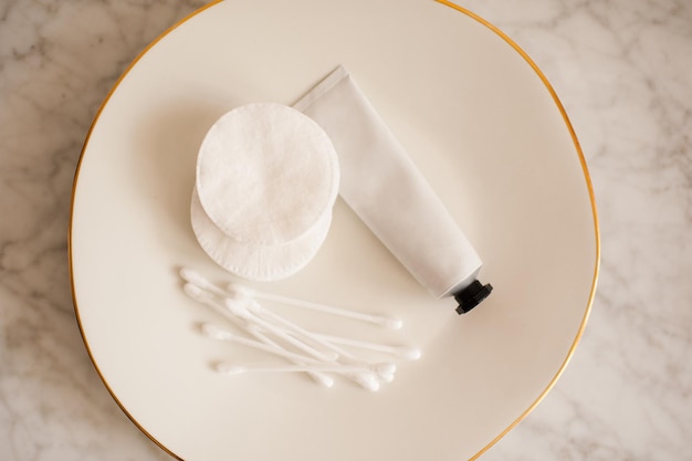 Mock up of hand cream package with cotton pads and sticks on white ceramic plate on table top view Beauty skincare products