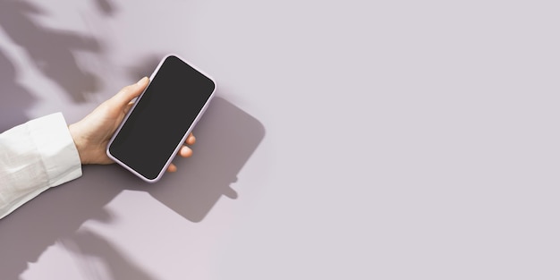 Mock up phone with black blank screen in female hand white shirt sleeve minimal flat lay on pastel violet background Summer lifestyle scene with smartphone sunlight shadows wide banner