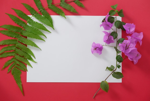 Mock-up white paper with space for text on red background and tropical leaves and flowers.