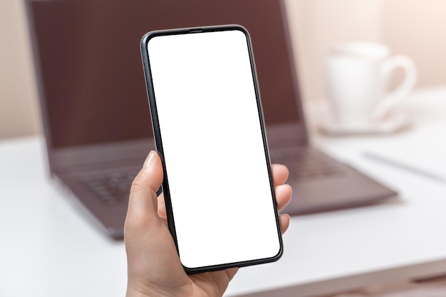 Mockup cellphone and laptop Woman hand holding phone with blank screen in office cell phone in woman's hand on background of laptop with white screen